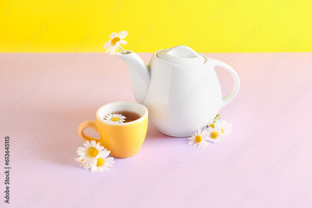 Teapot with cup of natural chamomile tea and flowers on pink table near yellow wall