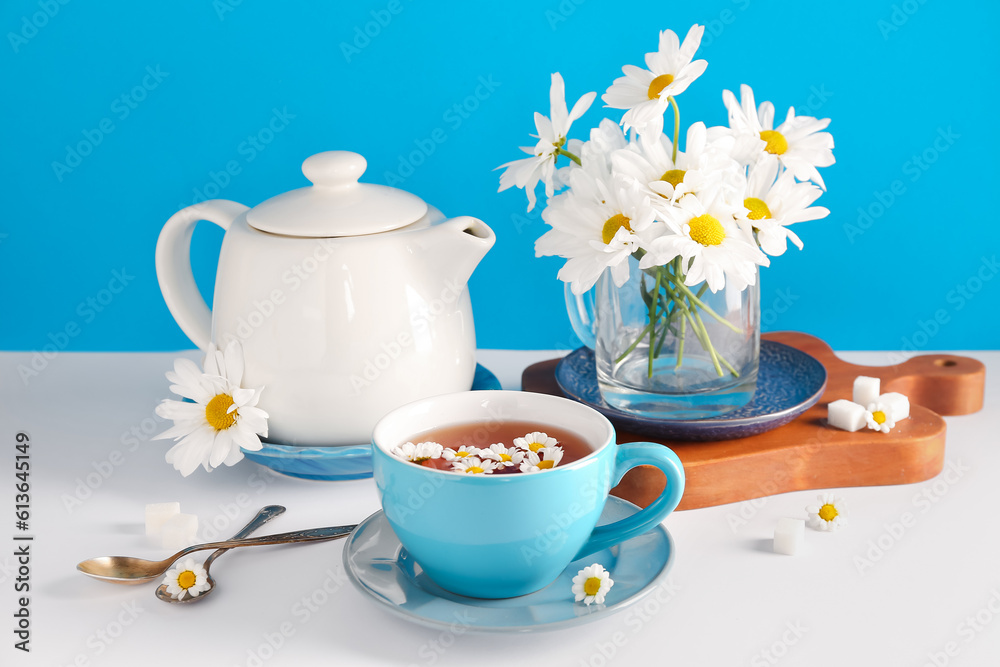 Wooden board with teapot, cup of natural chamomile tea and flowers on white table near blue wall