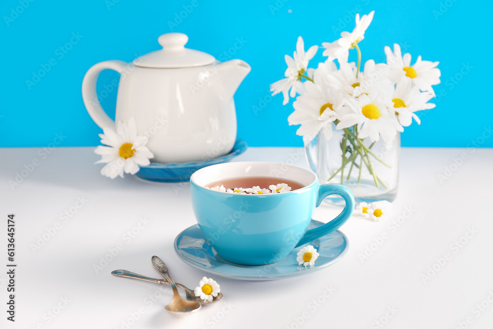 Teapot with cup of natural chamomile tea and flowers on white table near blue wall