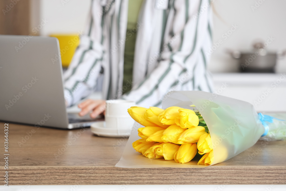 Woman using modern laptop at table with bouquet of yellow tulip flowers