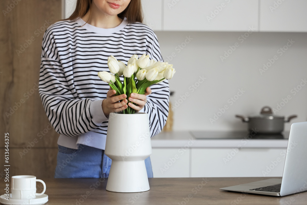 Woman putting bouquet of white tulip flowers into vase on wooden table in kitchen