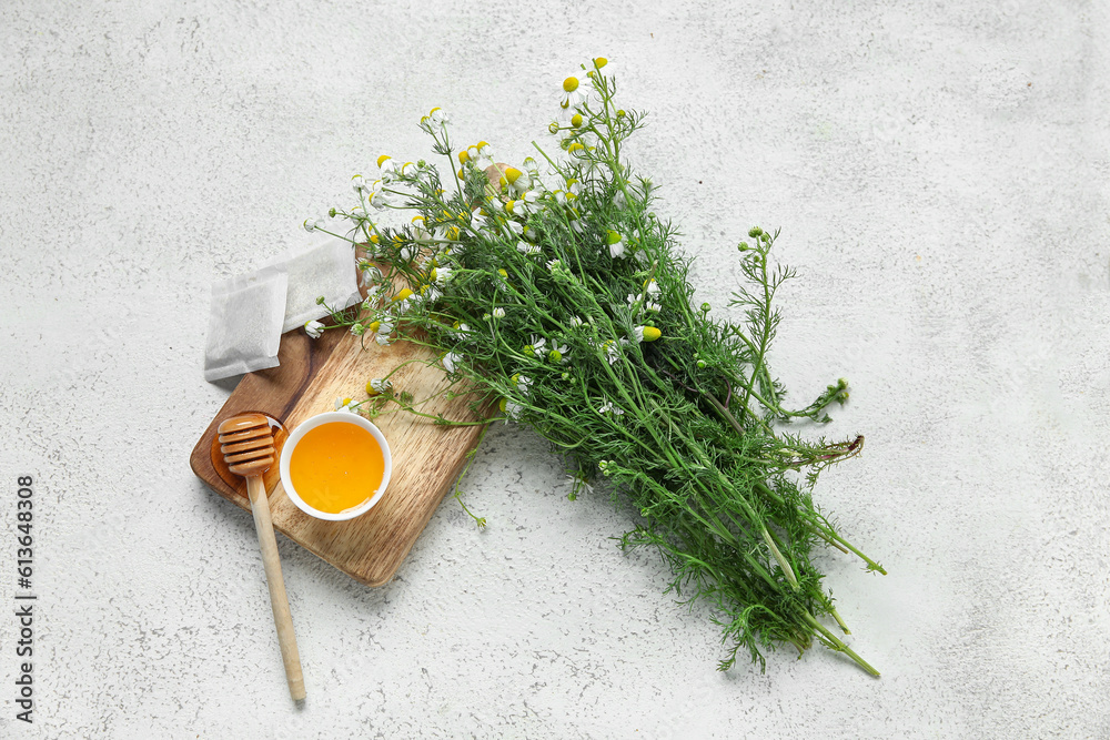 Wooden board with bowl of honey, tea bags and fresh chamomile flowers on light background