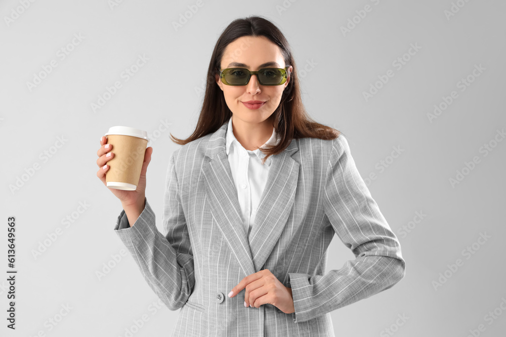 Young woman in stylish suit with cup of coffee on light background