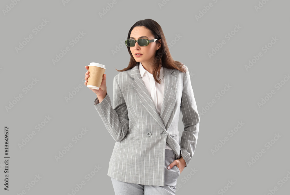 Young woman in stylish suit with cup of coffee on light background