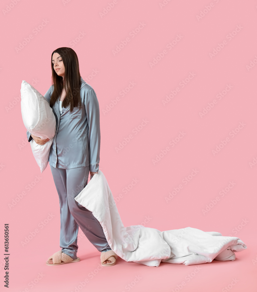 Young woman in pajamas with blanket and pillow on pink background