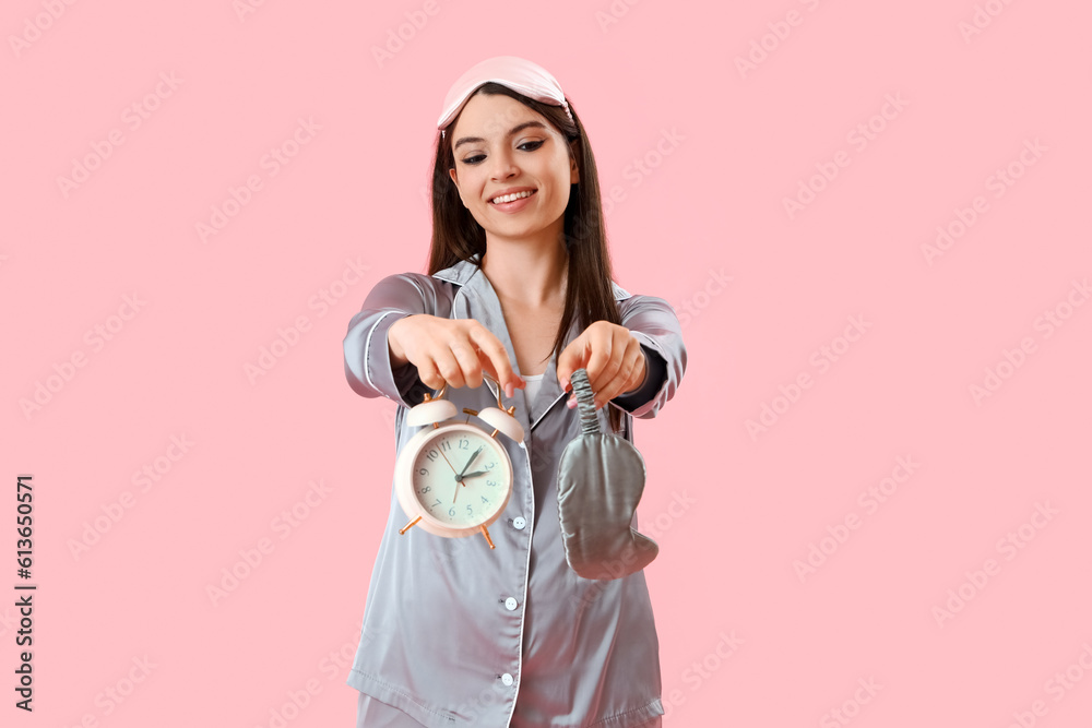 Young woman in pajamas with alarm clock and sleeping mask on pink background