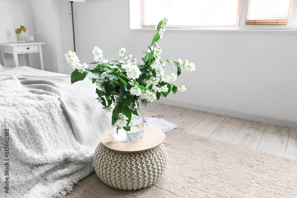 Vase with blooming jasmine flowers on pouf in interior of light bedroom