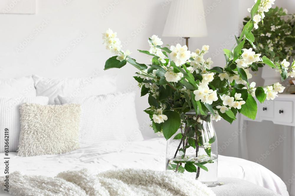 Vase with blooming jasmine flowers on bedside bench in interior of light bedroom