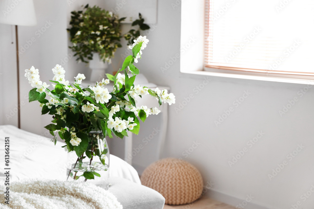 Vase with blooming jasmine flowers on bedside bench in interior of light bedroom