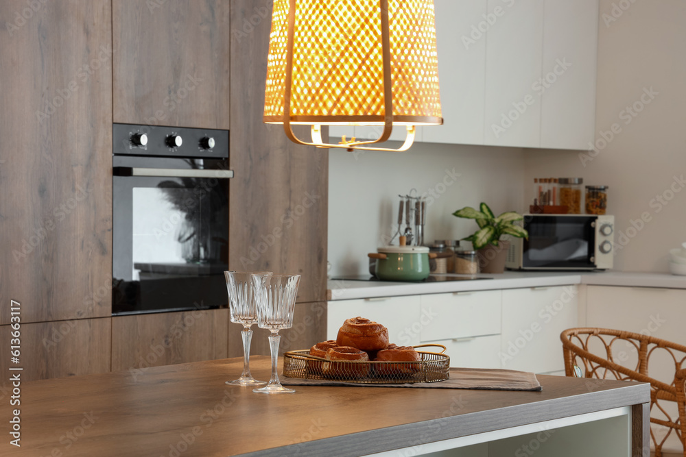 Wooden island table with delicious cinnamon rolls and wine glasses in modern kitchen at evening