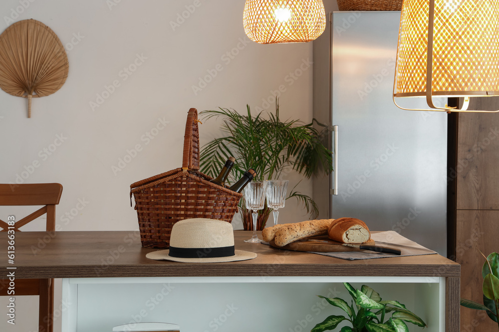 Hat, wicker basket and fresh bread on wooden island table in modern kitchen with glowing lamps