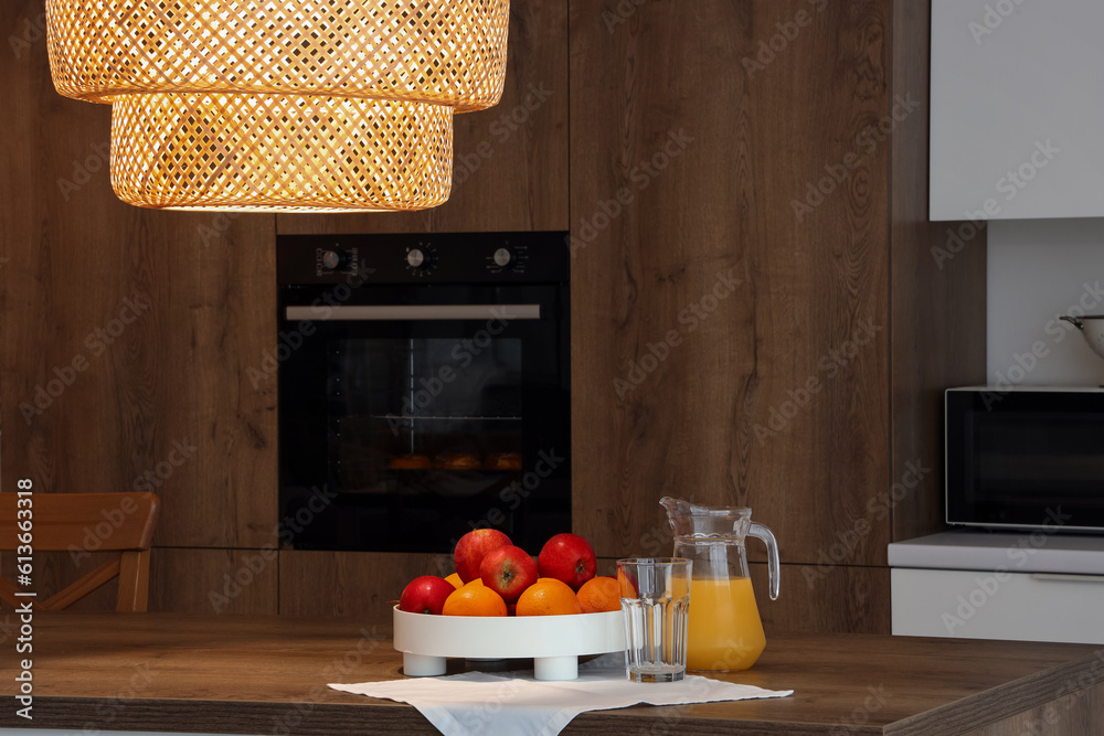 Jug with juice and fruits on wooden island table in modern kitchen in evening