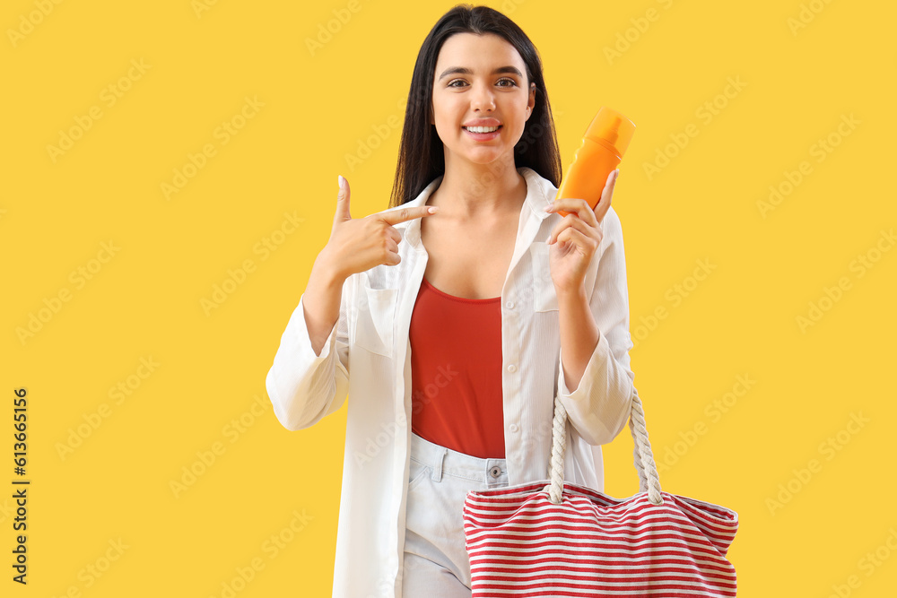 Young woman with beach bag pointing at sunscreen cream on yellow background