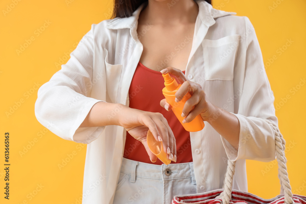 Young woman with beach bag applying sunscreen cream on yellow background, closeup