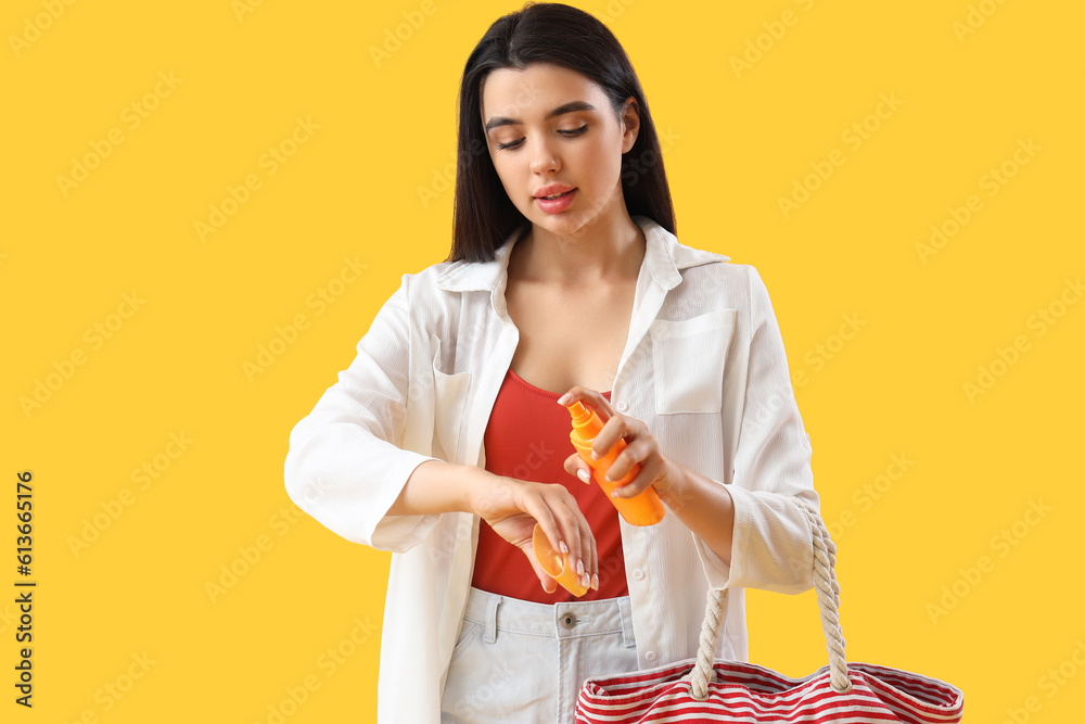 Young woman with beach bag applying sunscreen cream on yellow background