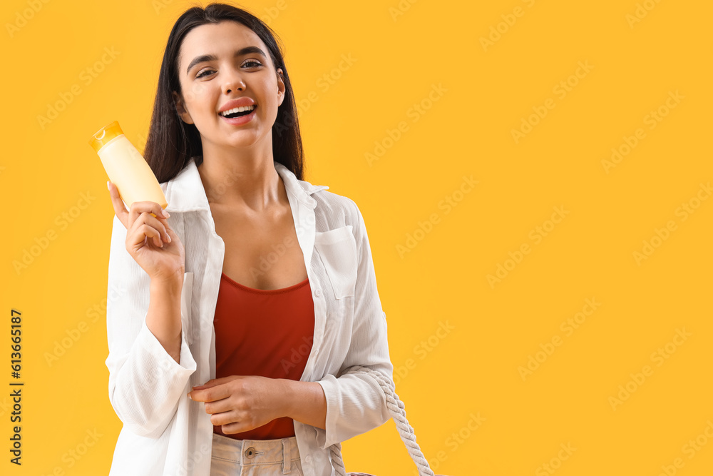 Young woman with sunscreen cream and beach bag on yellow background