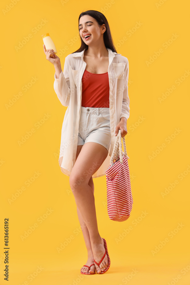 Young woman with sunscreen cream and beach bag on yellow background