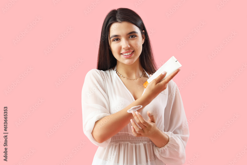 Young woman applying sunscreen cream on pink background