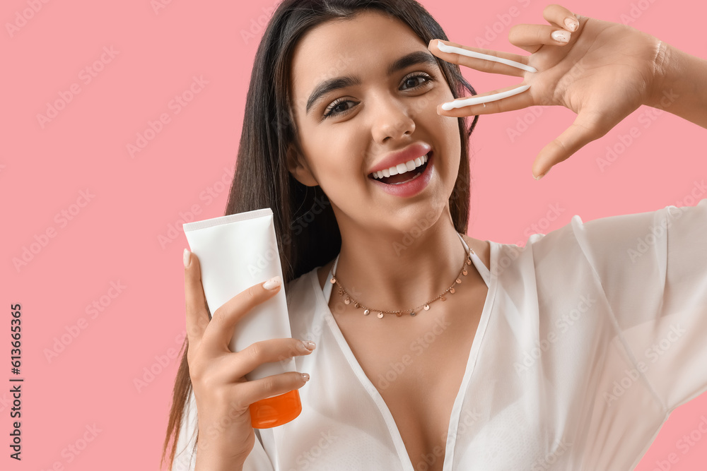 Young woman with sunscreen cream showing victory gesture on pink background