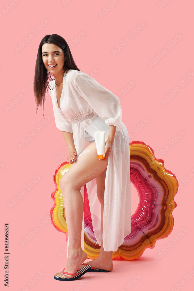 Young woman applying sunscreen cream onto her leg against pink background