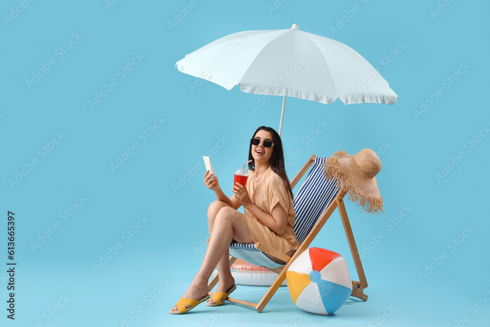 Young woman with sunscreen cream and soda in deck chair on blue background