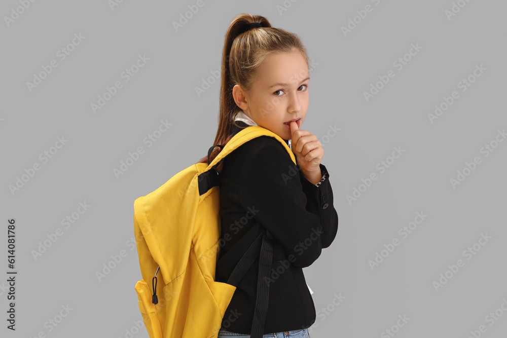 Little schoolgirl with backpack biting nails on grey background