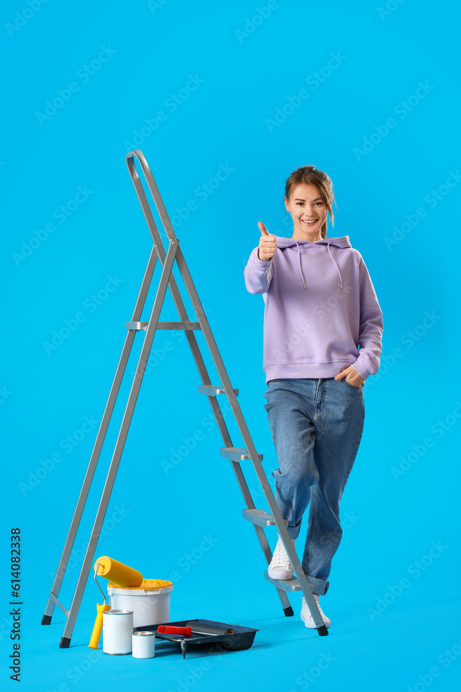 Young woman with ladder and paint cans showing thumb-up on blue background