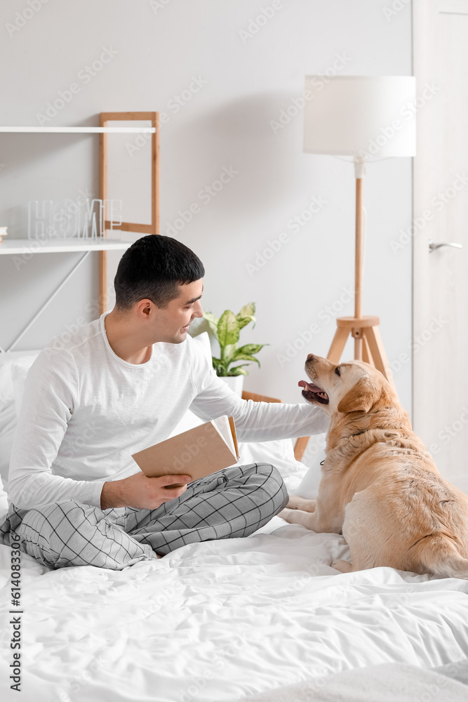 Young man with book and cute Labrador dog sitting in bedroom