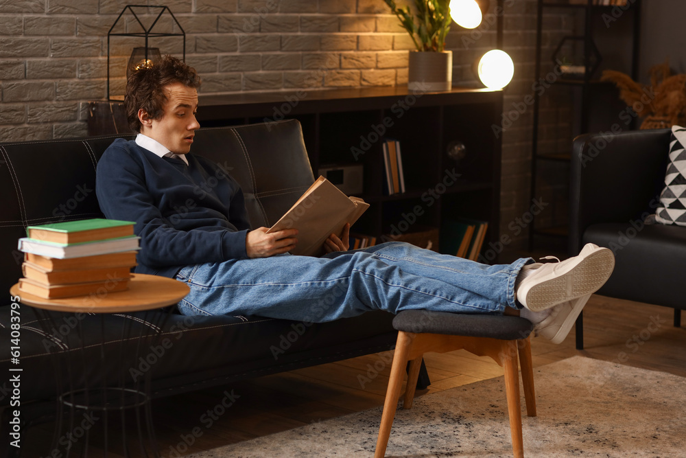 Young man reading book on sofa at home late in evening