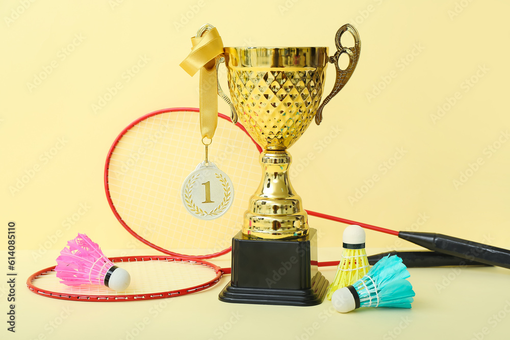 Gold cup with first place medal, badminton rackets and shuttlecocks on yellow background