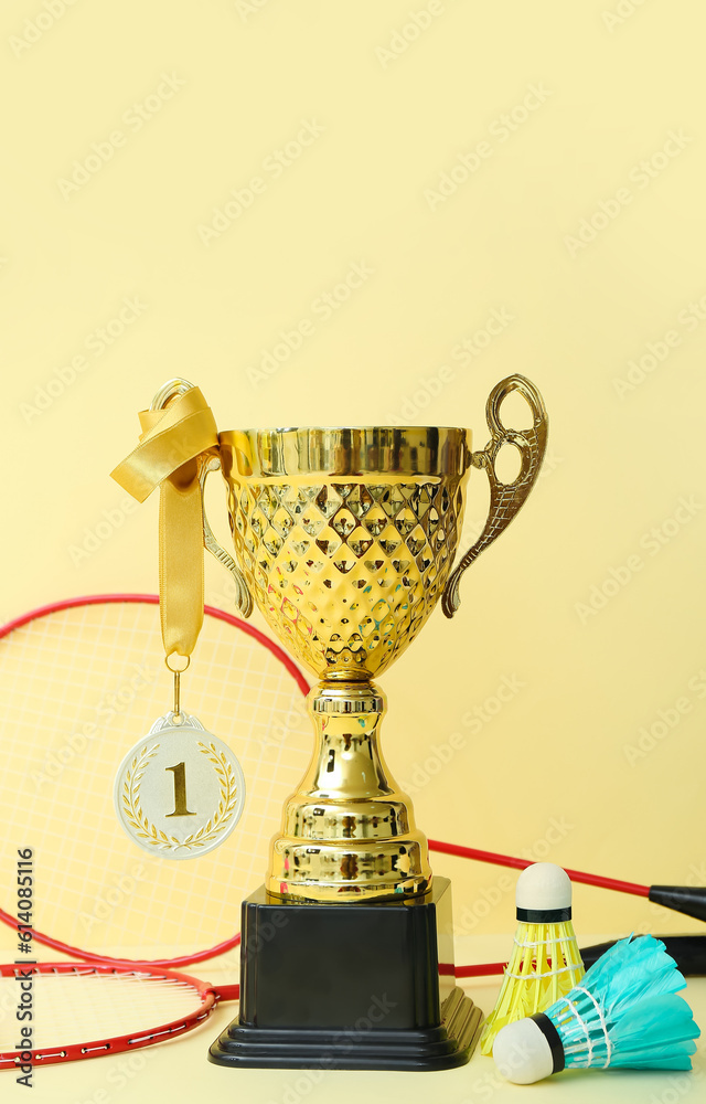 Gold cup with first place medal, badminton rackets and shuttlecocks on yellow background