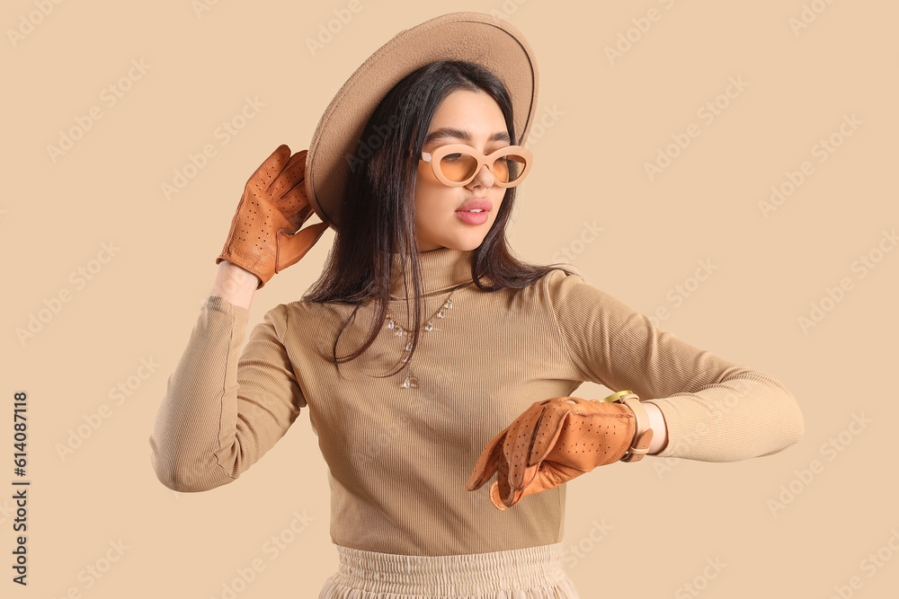 Stylish young woman in leather gloves looking at wristwatch on beige background