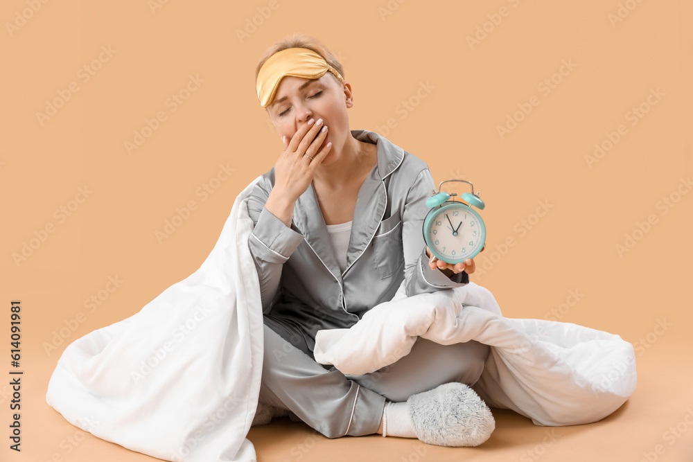 Yawning young woman with alarm clock and blanket on beige background