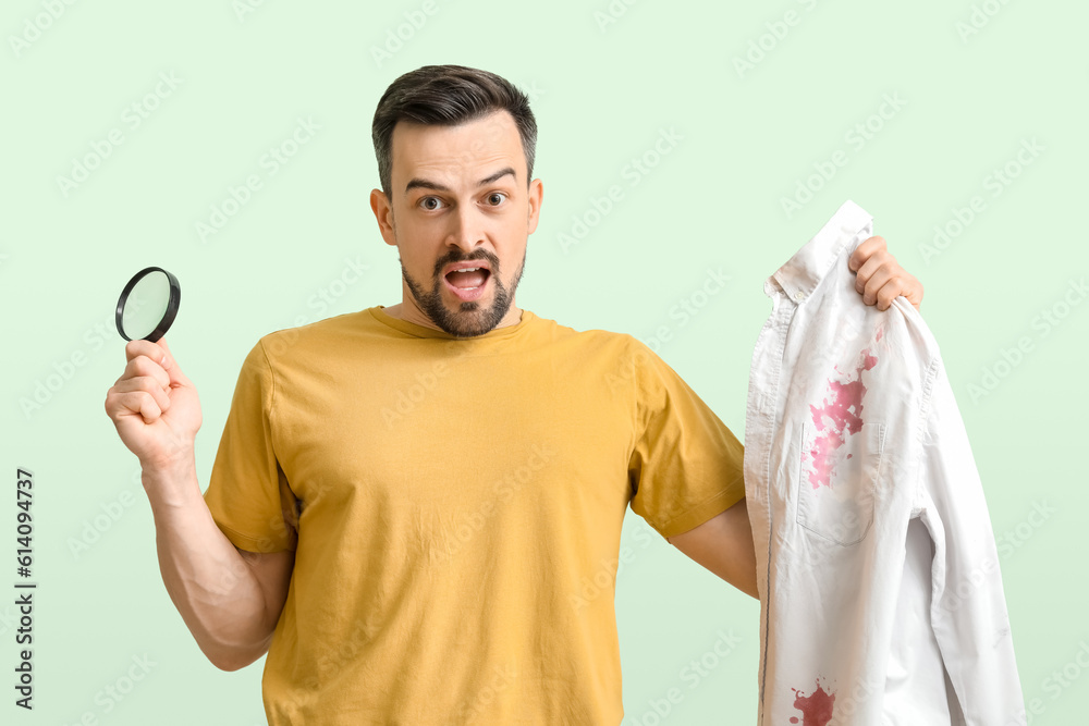 Shocked young man with magnifier and stained shirt on green background