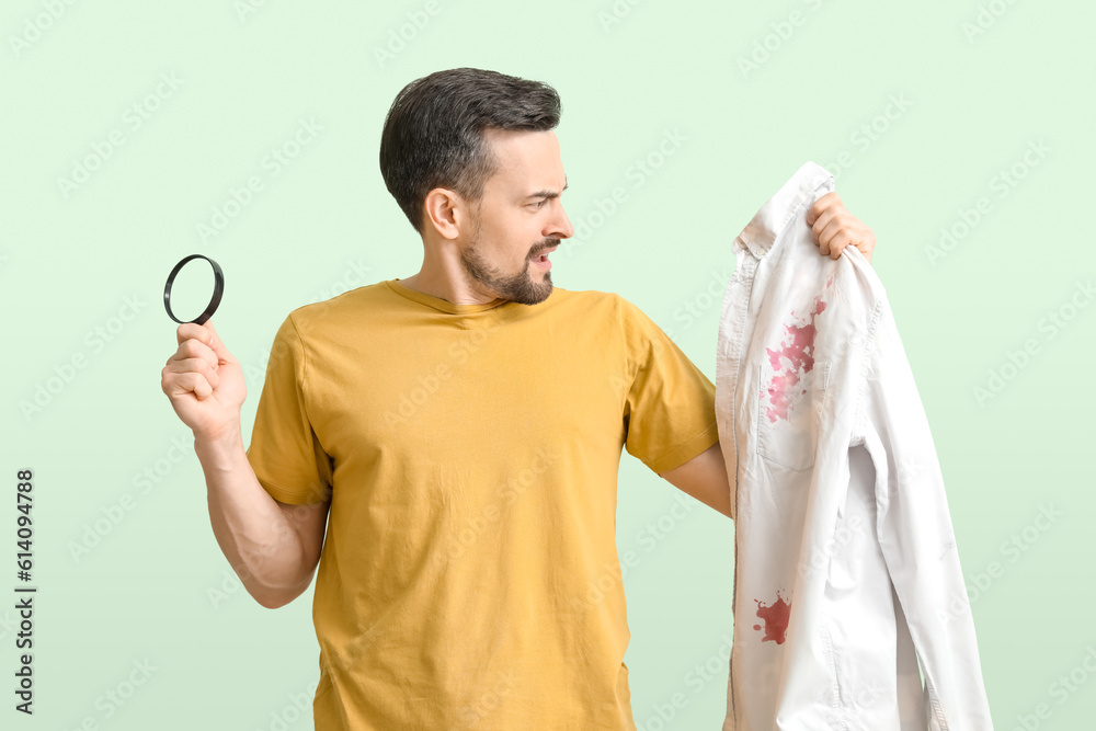 Shocked young man with magnifier and stained shirt on green background