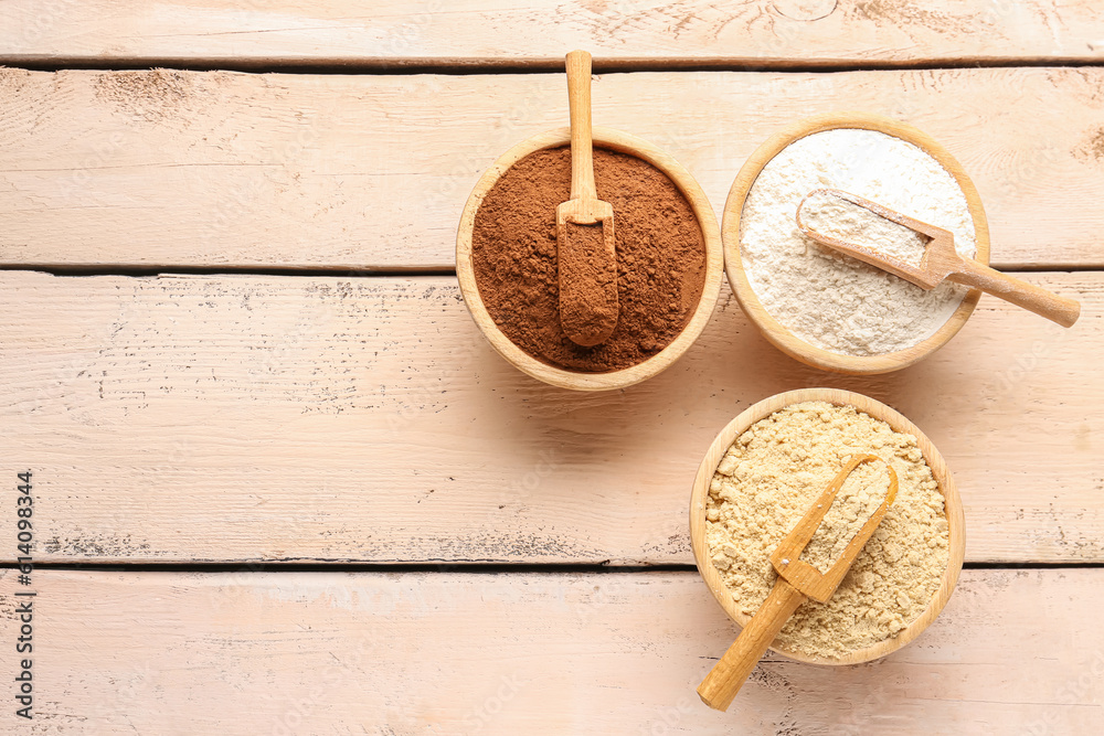 Wooden bowls with different types of flour and cacao on light wooden background