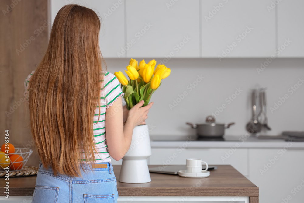 Beautiful young woman putting bouquet of yellow tulip flowers into vase in modern kitchen