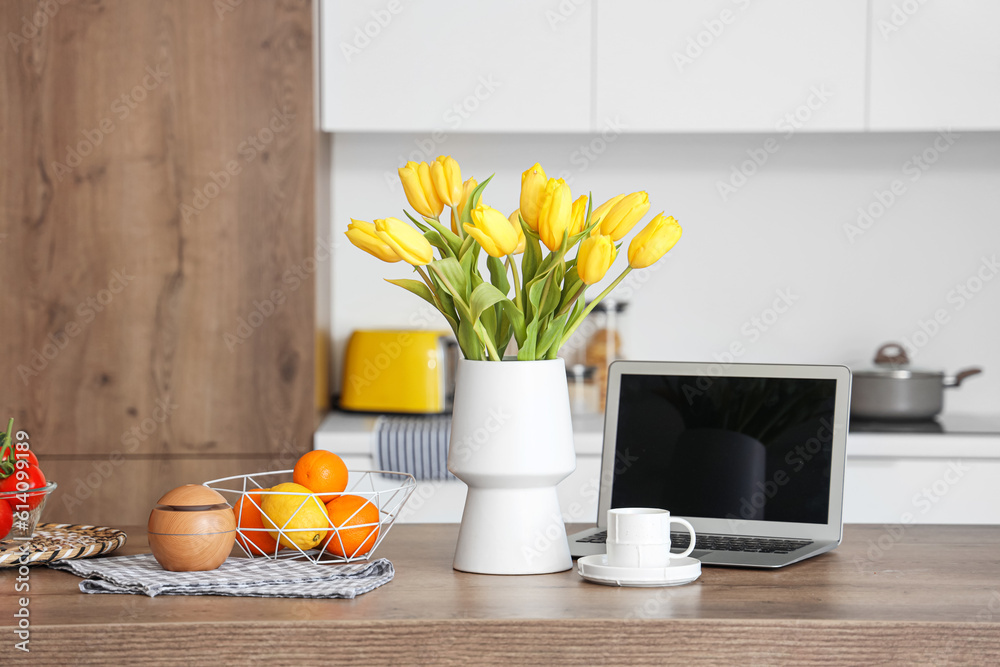 Vase with yellow tulip flowers, modern laptop, fruits and cup of coffee on wooden table in light kit