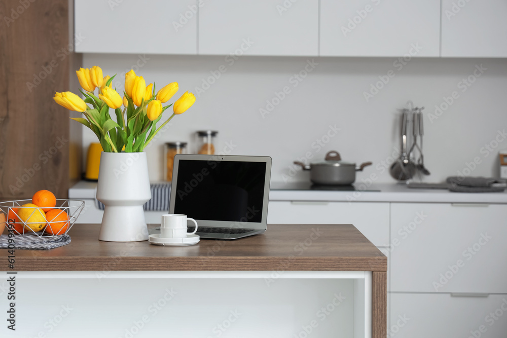 Vase with yellow tulip flowers, modern laptop, fruits and cup of coffee on wooden table in light kit