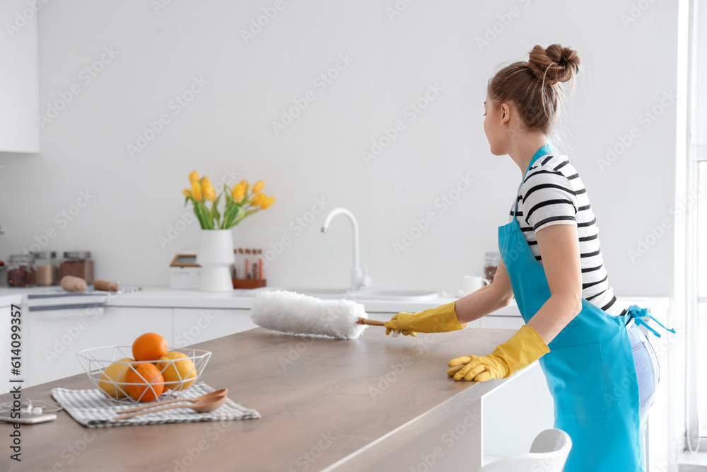 Pretty young woman cleaning dining table with dust brush in light kitchen