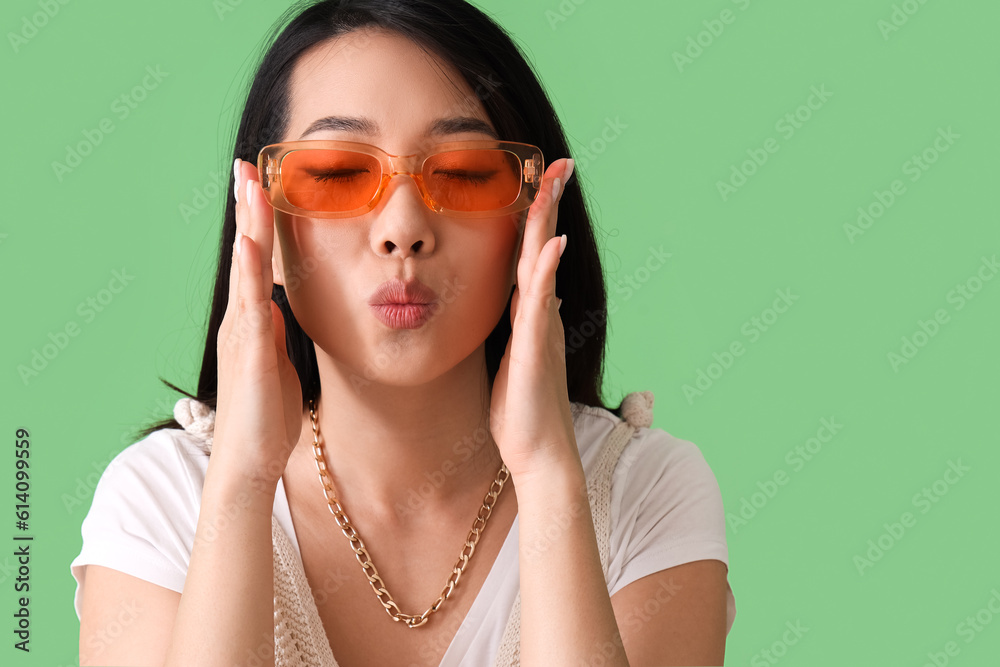 Beautiful Asian woman in stylish sunglasses blowing kiss on green background, closeup