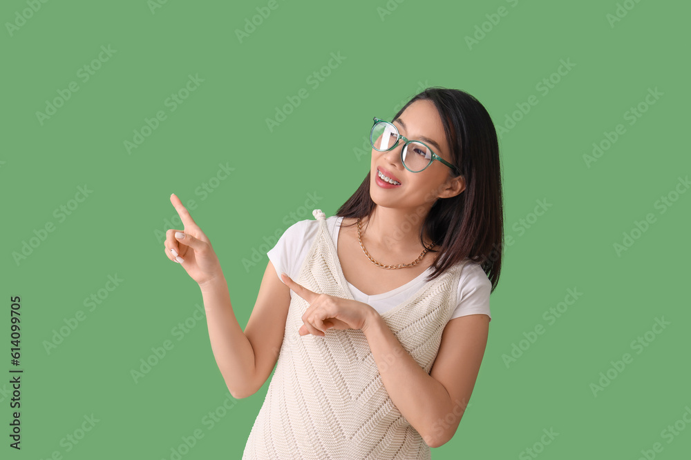 Beautiful Asian woman in stylish eyeglasses pointing at something on green background