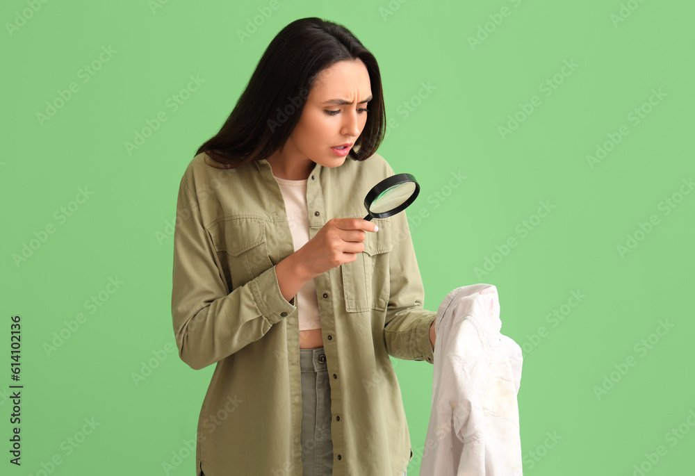 Young woman with magnifier and stained clothes on green background