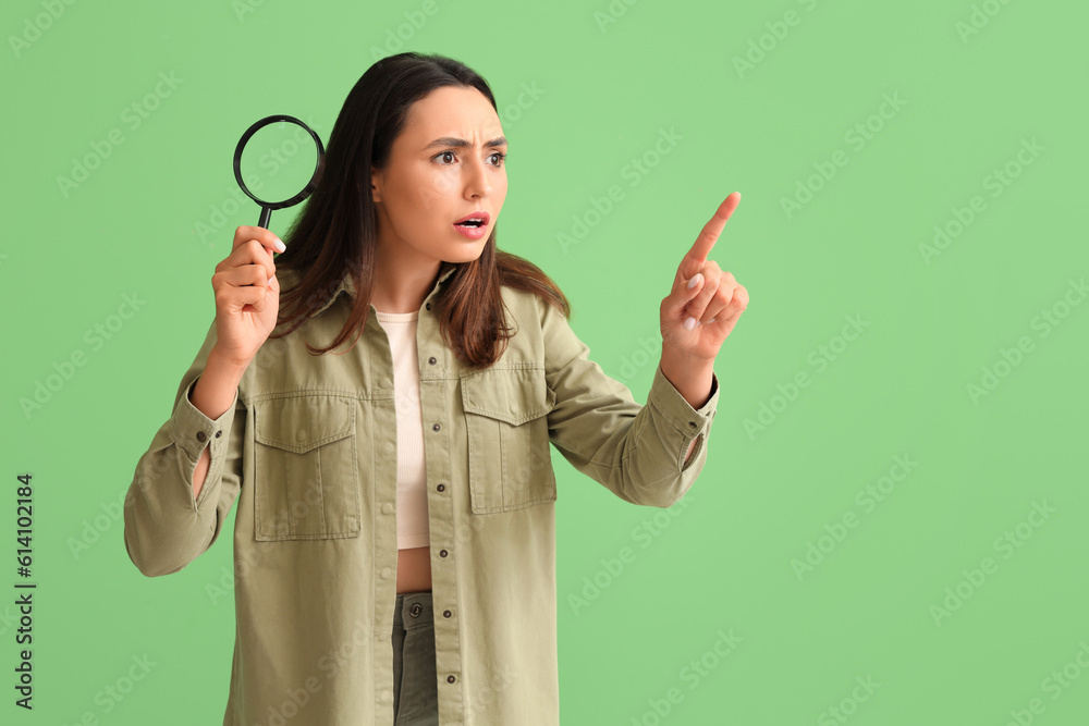 Shocked young woman with magnifier pointing at something on green background