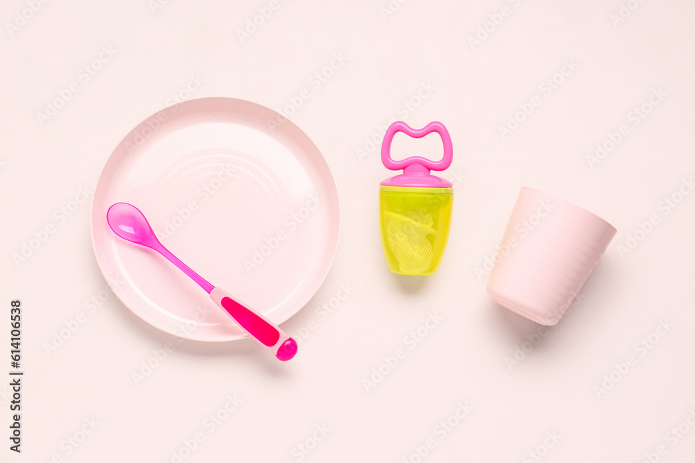 Plate with spoon, cup and nibbler for baby on pink background