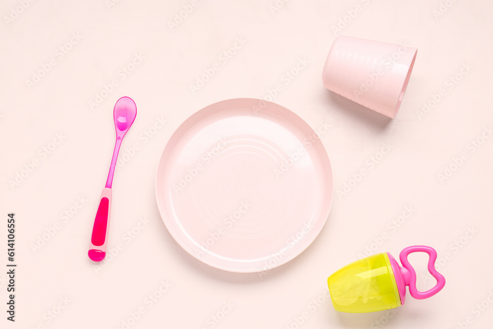 Plate with spoon, cup and nibbler for baby on pink background