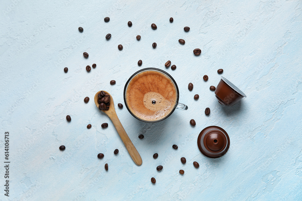 Cup of delicious coffee, wooden spoon, beans and capsules on blue table