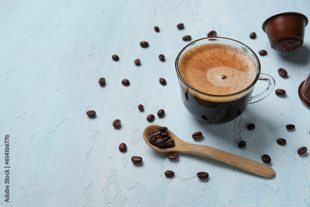 Cup of delicious coffee, wooden spoon, beans and capsule on blue table