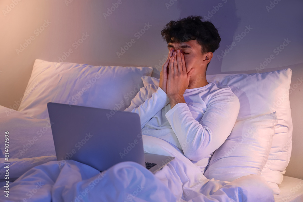 Sleepy young man using laptop in bed at night