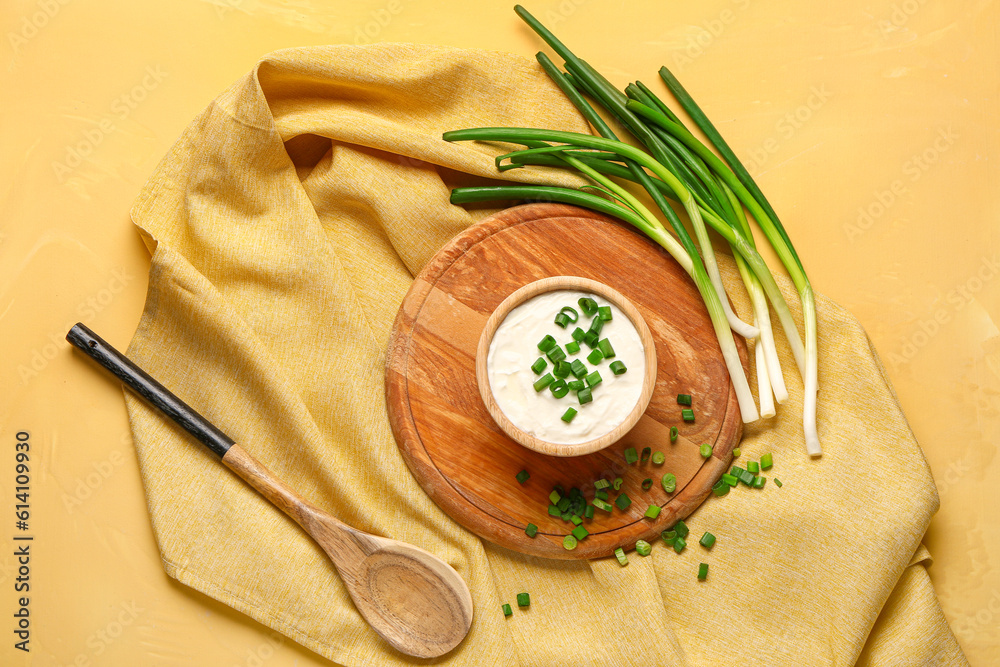 Board with bowl of tasty sour cream and sliced green onion on yellow background